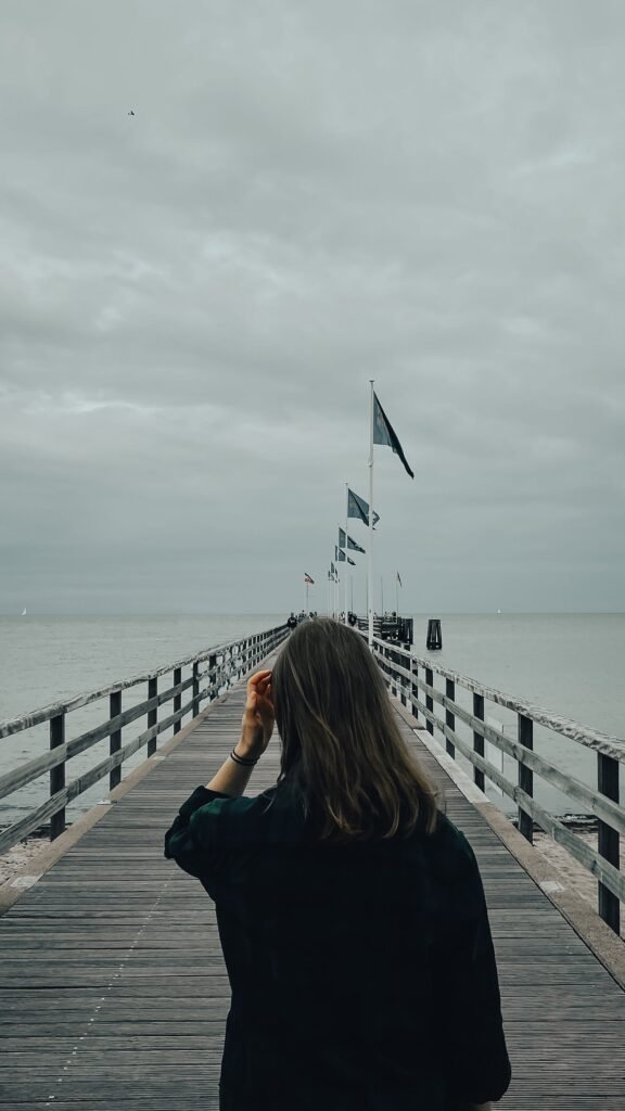 A person walks on a wooden pier extending into a calm sea under a cloudy sky.